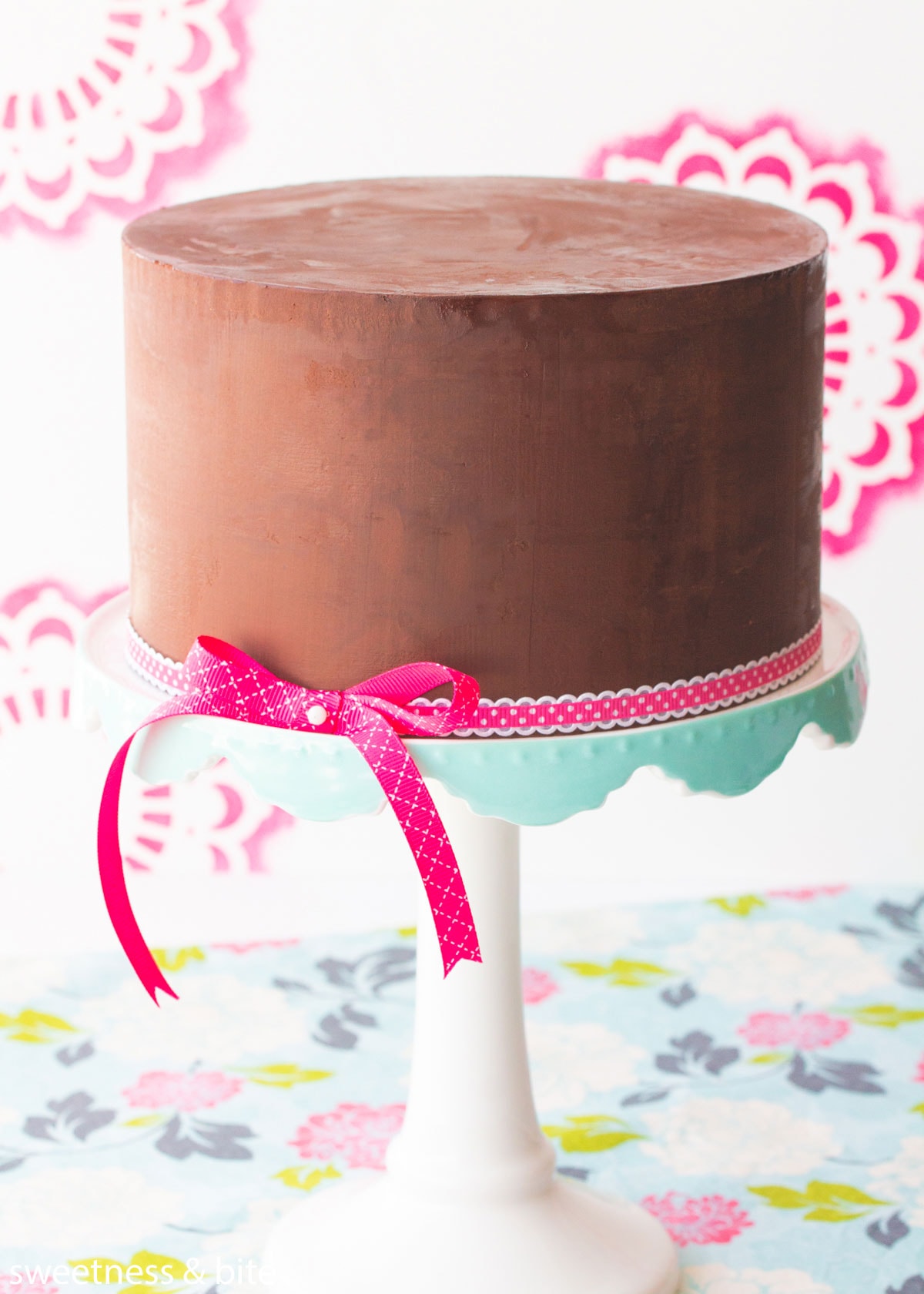 A dark chocolate ganache covered cake, sitting on a white and blue scalloped cake stand, wrapped with a pink and white ribbon tied in a bow at the base.