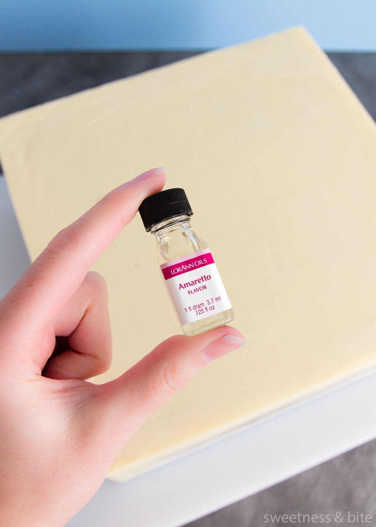 A hand holding a small bottle of Lorann Amaretto flavouring over a white chocolate ganache covered square cake.