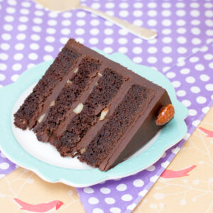 A slice of chocolate cake, filled with ganache and chopped almonds, on a green scalloped plate, sitting on a purple spotted cloth.