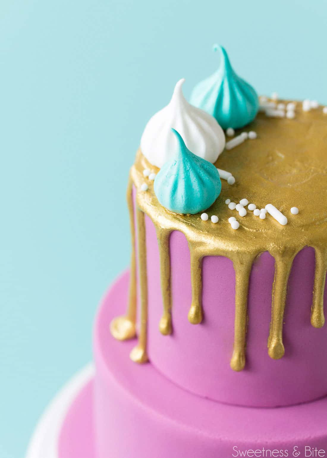 Close up of the top tier of the mini cake, with the gold drip and decorated with white and teal mini meringues and white sprinkles.