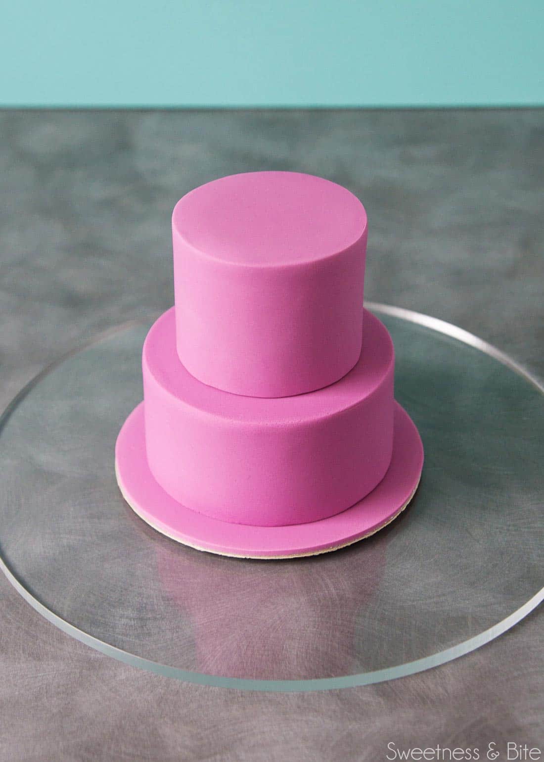 The two tiers of cake covered in purple fondant, stacked on the fondant covered cake board.