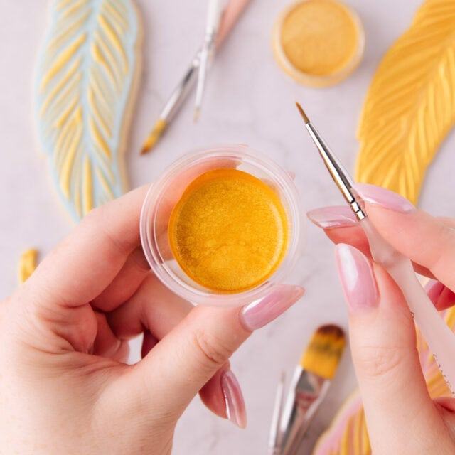 A close up of two hands with light pink nail polish, one is holding a small container of edible gold paint, the other is holding a fine paintbrush with a white handle, and there are gold painted cookies in the background.