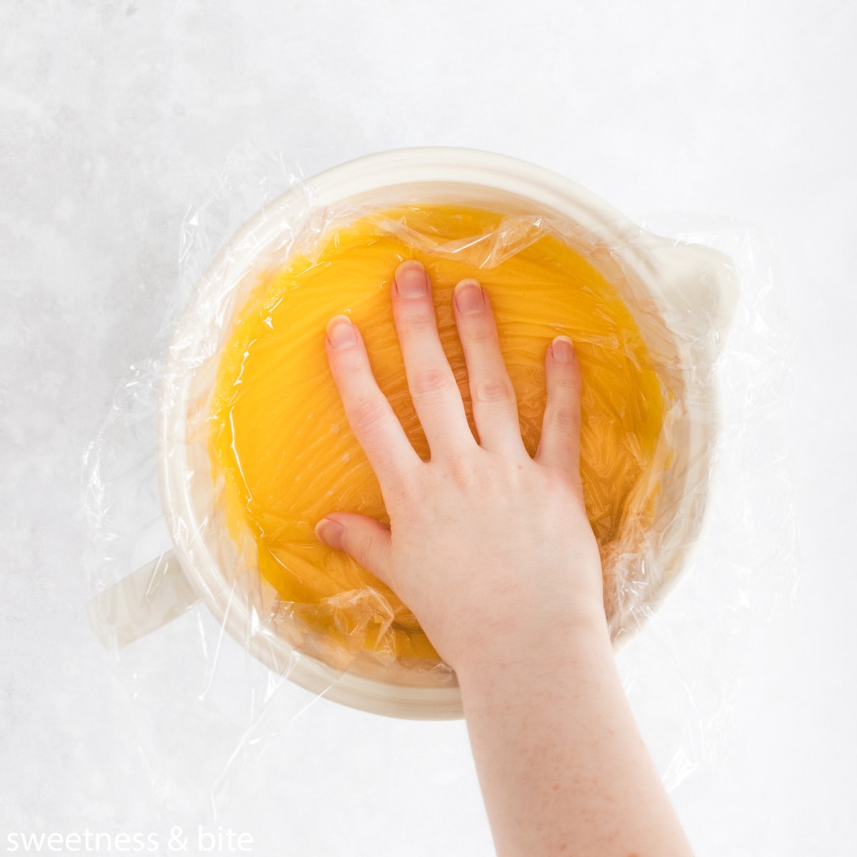 Lemon curd in a large jug, with a hand pressing plastic wrap onto the surface.