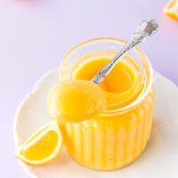 Microwave lemon curd in a ribbed glass jar, with a silver spoon on a purple background.