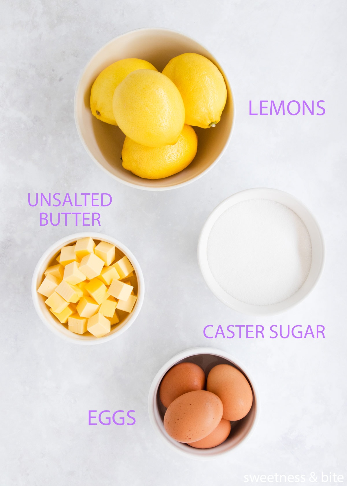The lemon curd ingredients in white bowls on a grey background - lemons, butter, sugar and eggs.