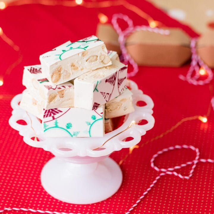 Pieces of almond and honey nougat in a white glass bowl on a festive red background.