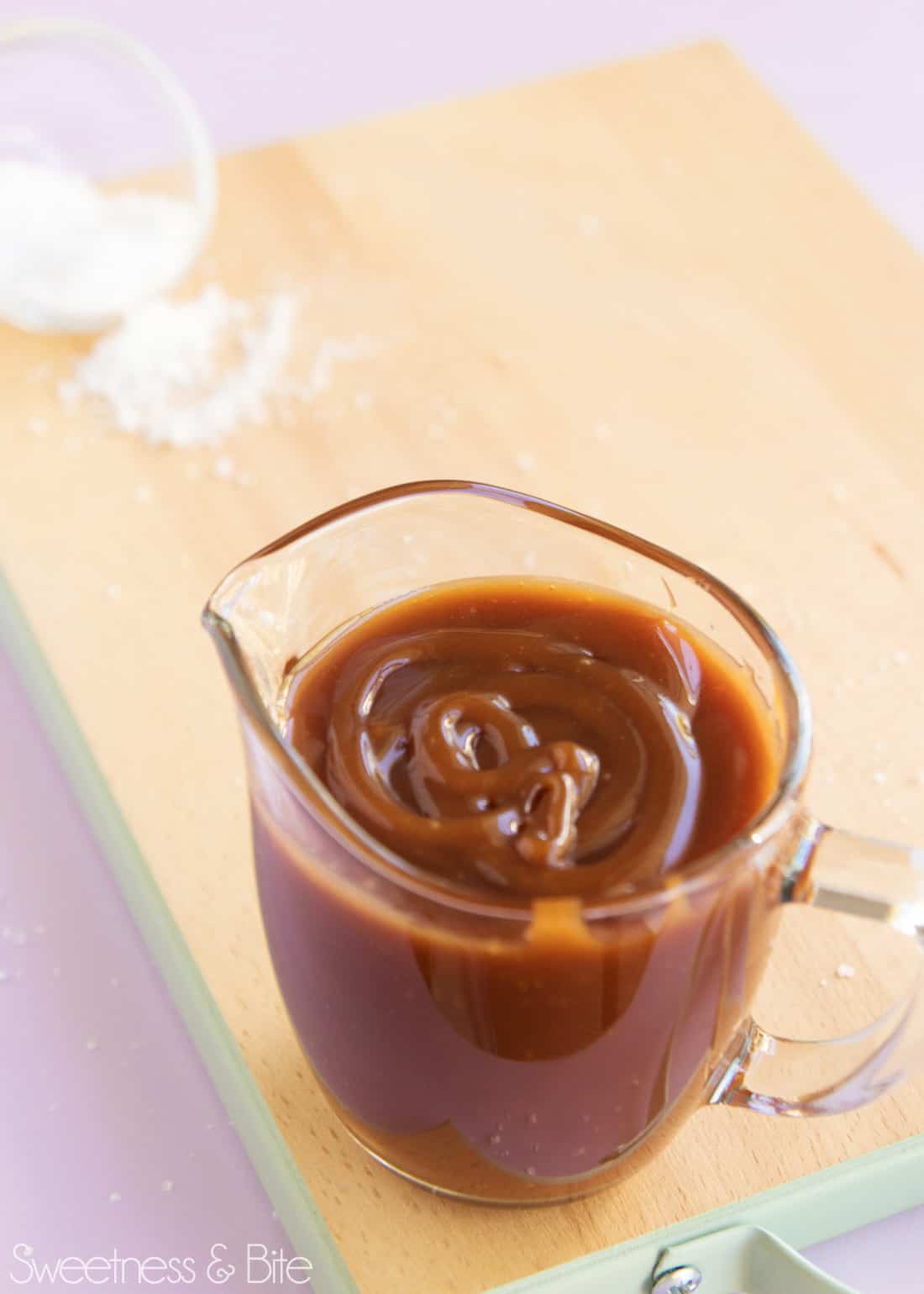 A small glass jug full of caramel sauce, on a light wooden chopping board sprinkled with flaky salt.