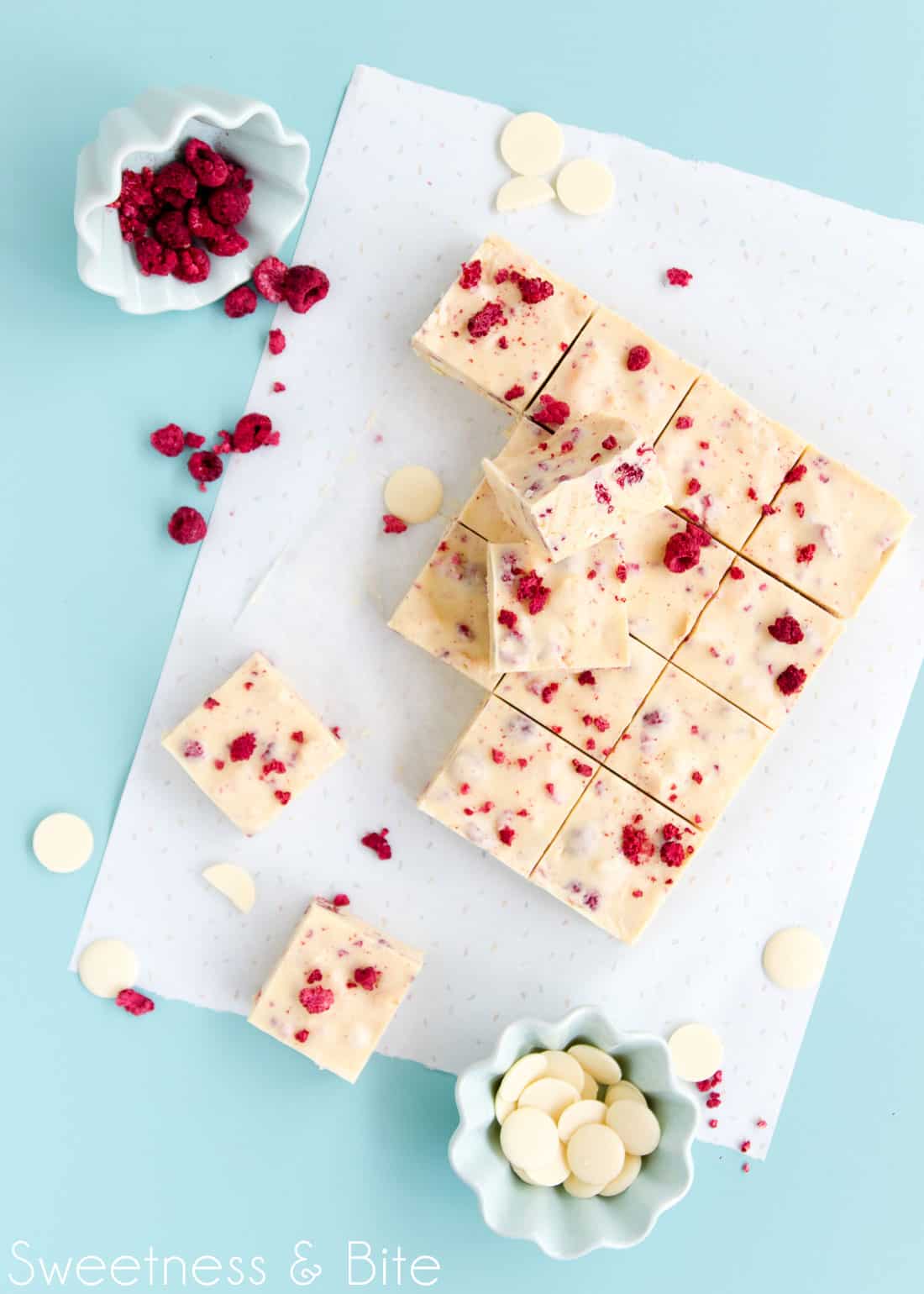 Two pieces of slice, sitting on top of the remaining pieces of slice below. A small green bowl of freeze-dried raspberry pieces sits on its side, with scattered pieces of raspberry falling out. White chocolate buttons and broken freeze-dried raspberries are scattered across the blue-green tabletop.