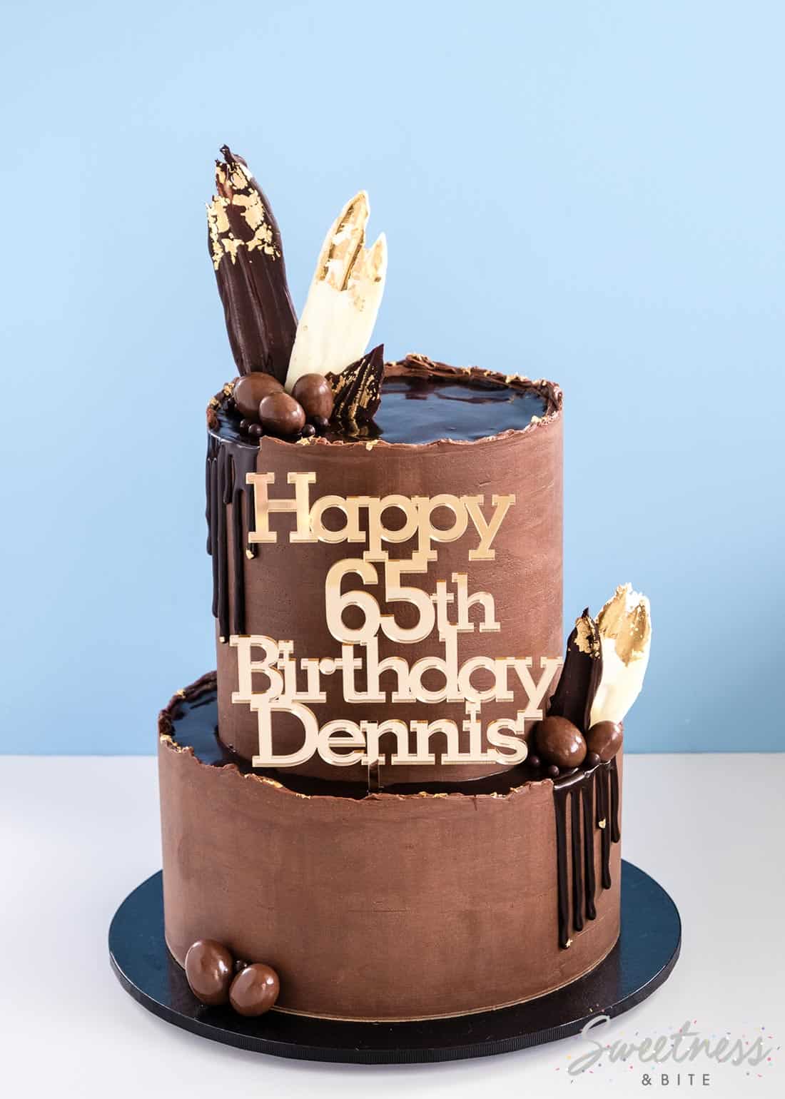 A two tier chocolate ganache covered cake with ganache drips and a gold happy birthday cake topper.