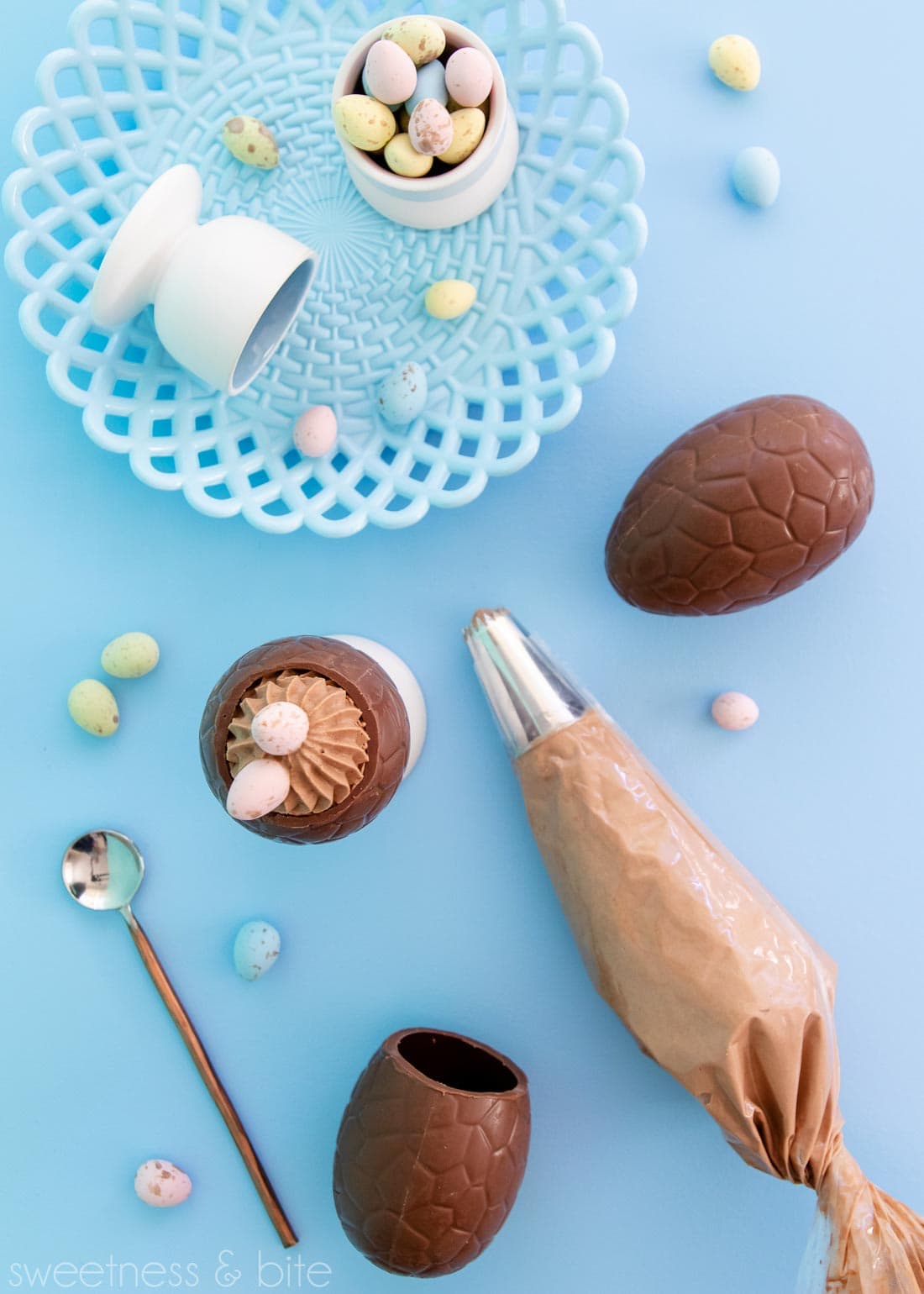 Flat lay including a mousse filled egg, piping bag full of mousse, a blue plate and pastel mini eggs, on a blue background.