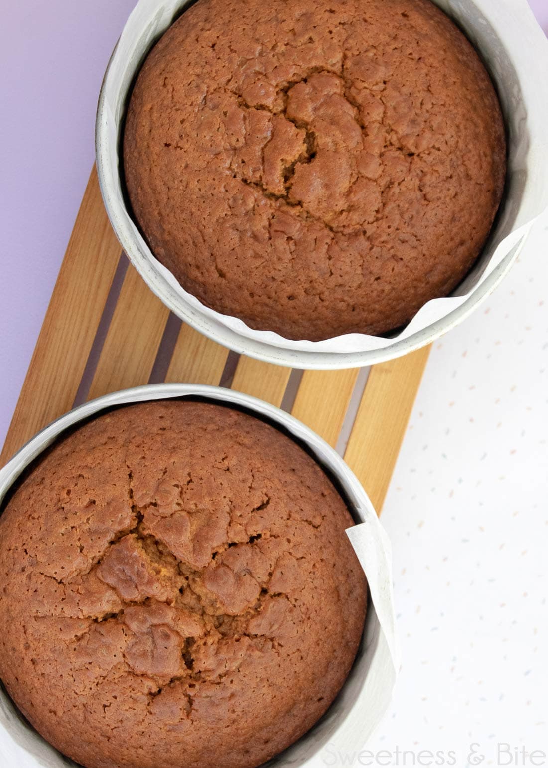 Two cakes in cake pans on a wooden cooling rack.