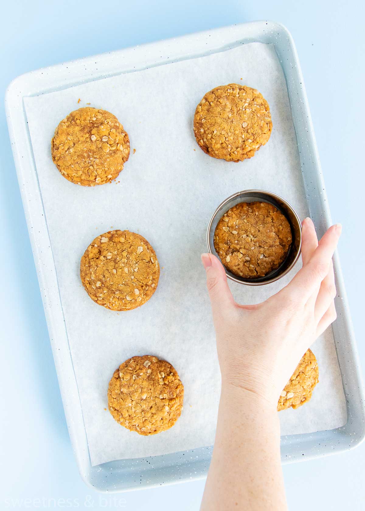 Six baked Anzac biscuits on a tray, with a round cookie cutter being used to make the biscuits more round.