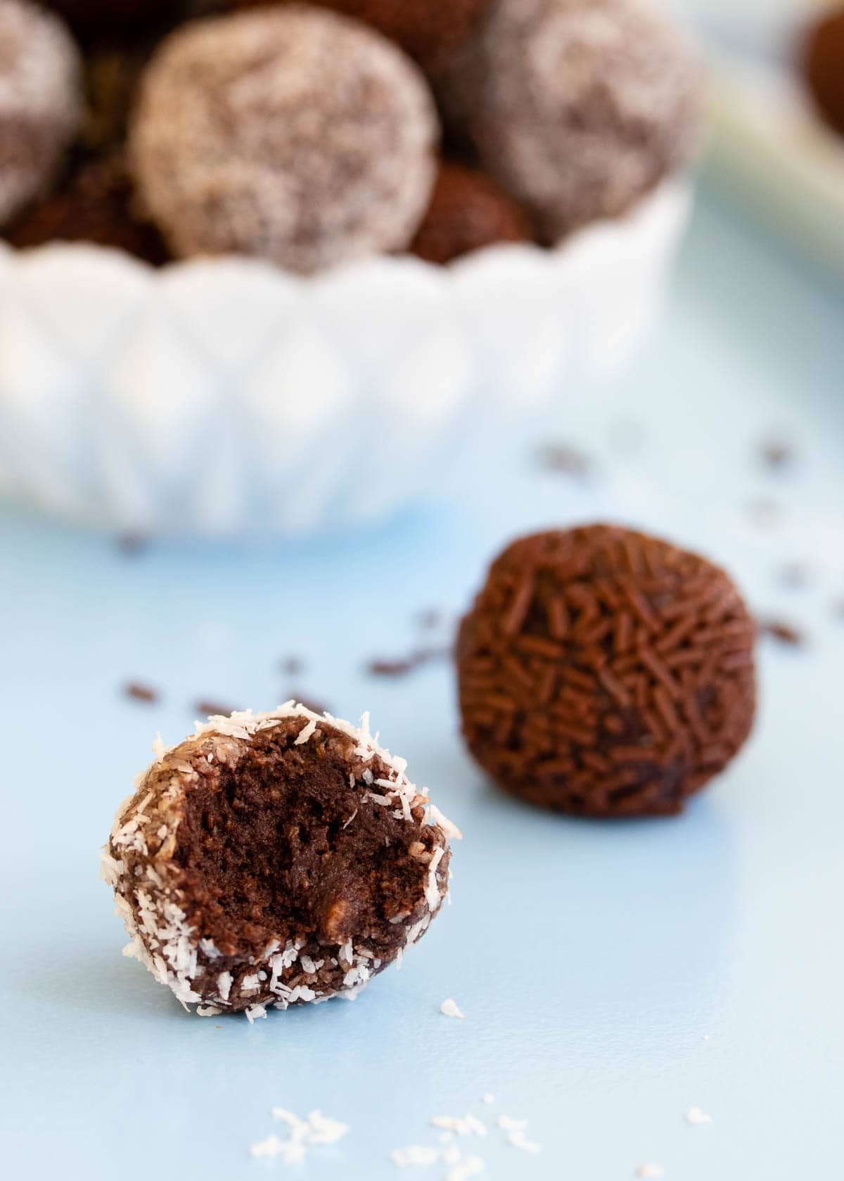 A chocolate coconut truffle with a bite taken out of it, on a bale blue background with a bowl of truffles in the distance.