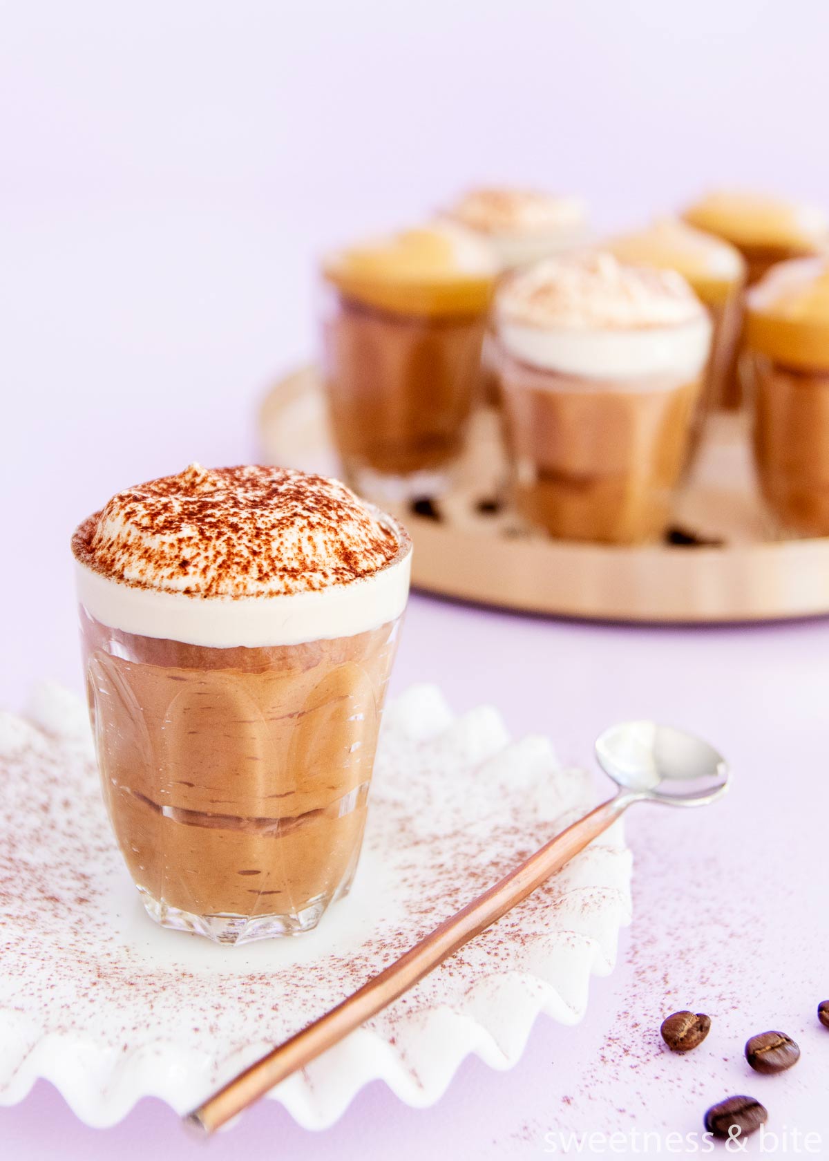 A small glass of mocha mousse with whipped cream topping dusted with chocolate powder, on a white milk glass plate with a ruffled edge, and a copped handled spoon.