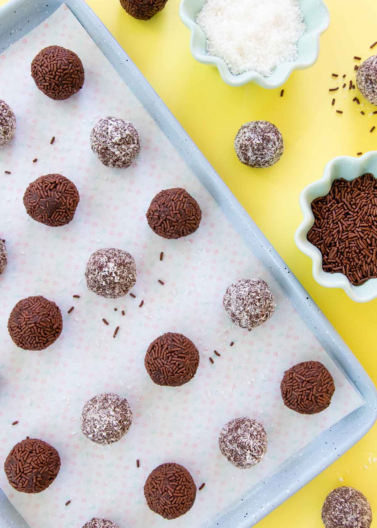 Chocolate coconut truffles on a light blue baking tray, with small bowls of chocolate sprinkles and desiccated coconut.