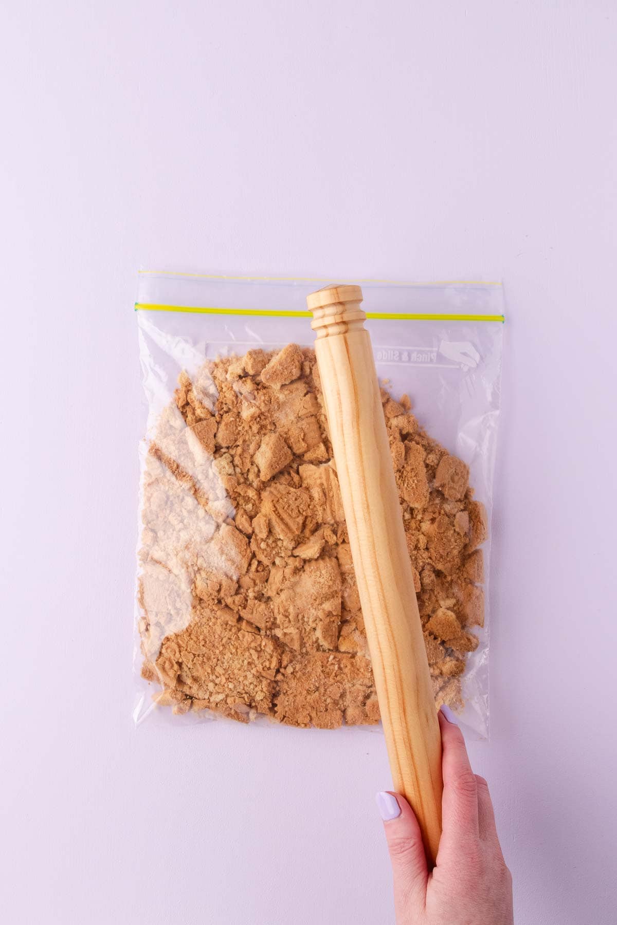Plain biscuits in a resealable plastic bag, being crushed into small pieces with a wooden rolling pin.