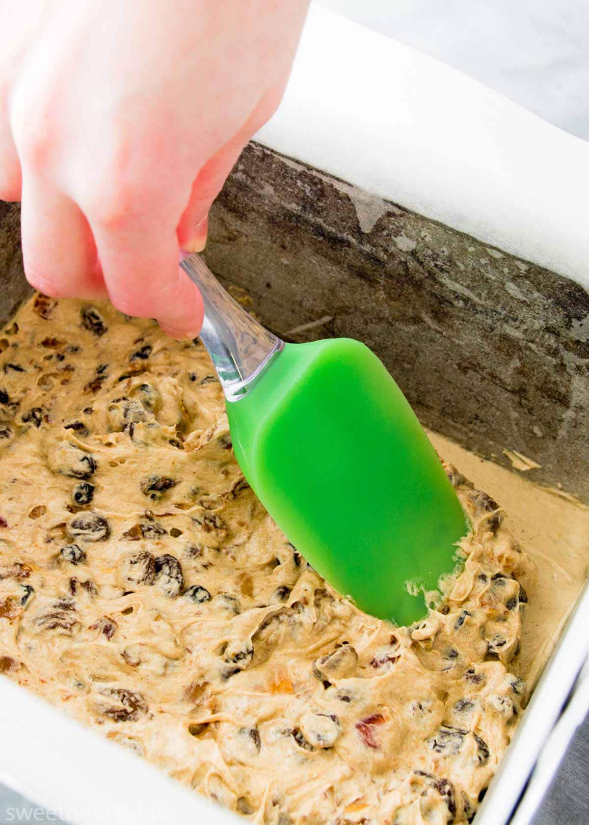 Fruit cake batter being pressed into the prepared cake pan with the back of a spatula.