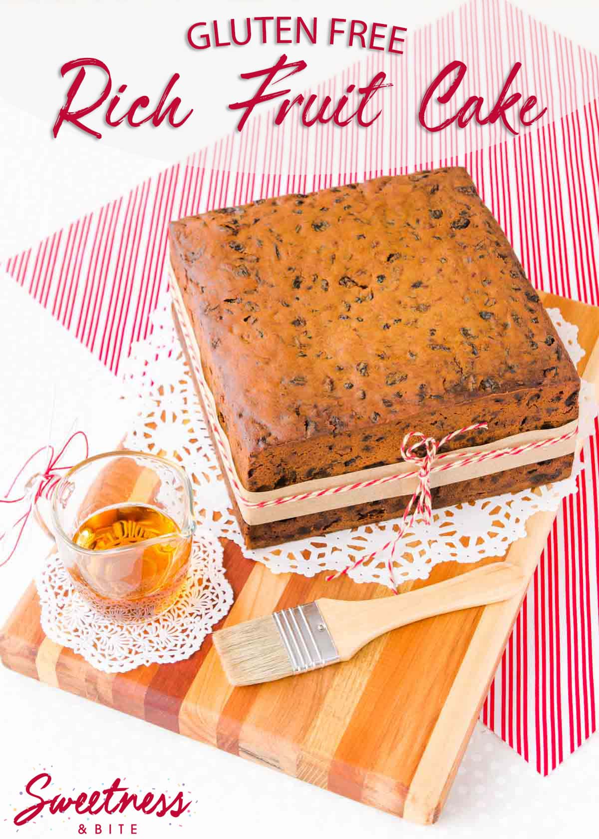 Square gluten free fruit cake on a wooden board, sitting on a red and white striped cloth, with a glass jug of brandy and a pastry brush.