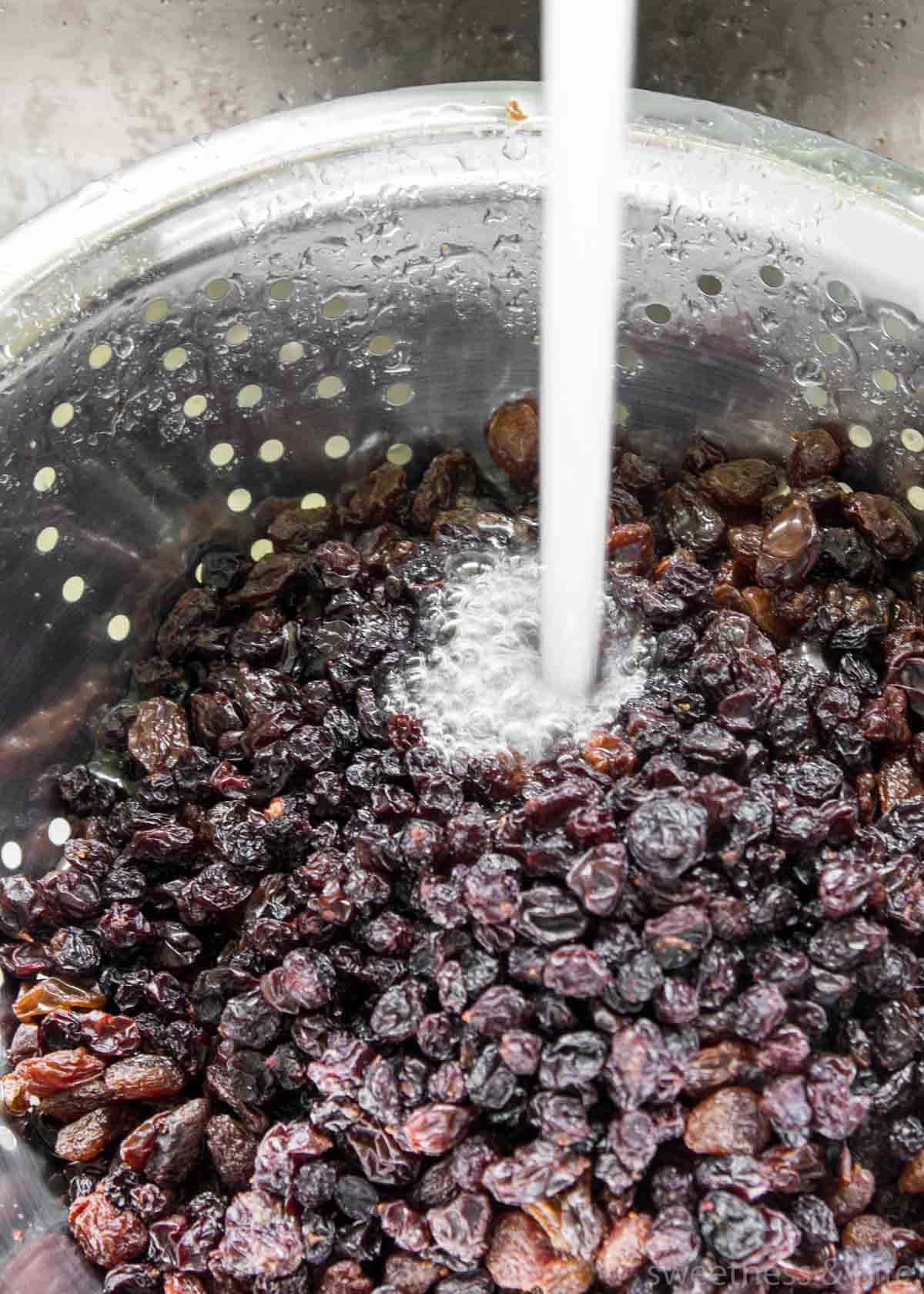 Raisins being rinsed under a tap in a metal colander.