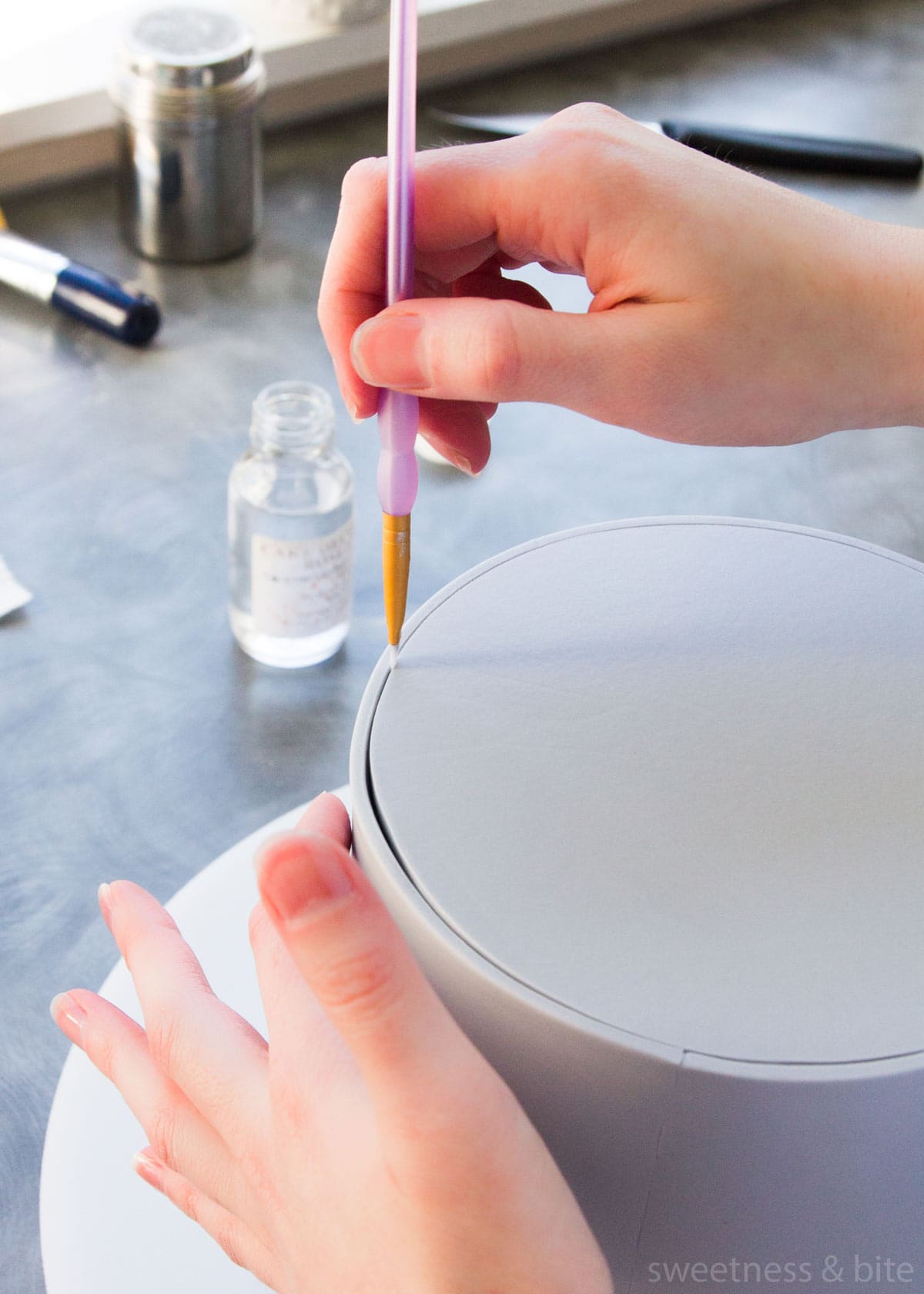 A round cake wrapped in grey fondant, with a small paintbrush being used to paint vodka into the join to attach the fondant.