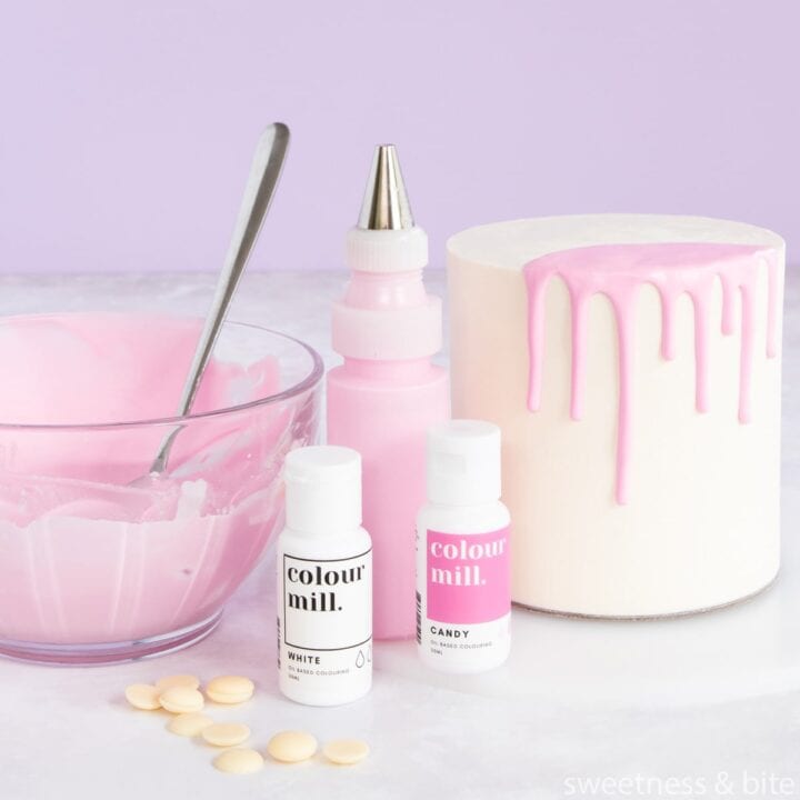 A white cake with a pink coloured ganached drip, a bowl of pink ganache and food colouring bottles.