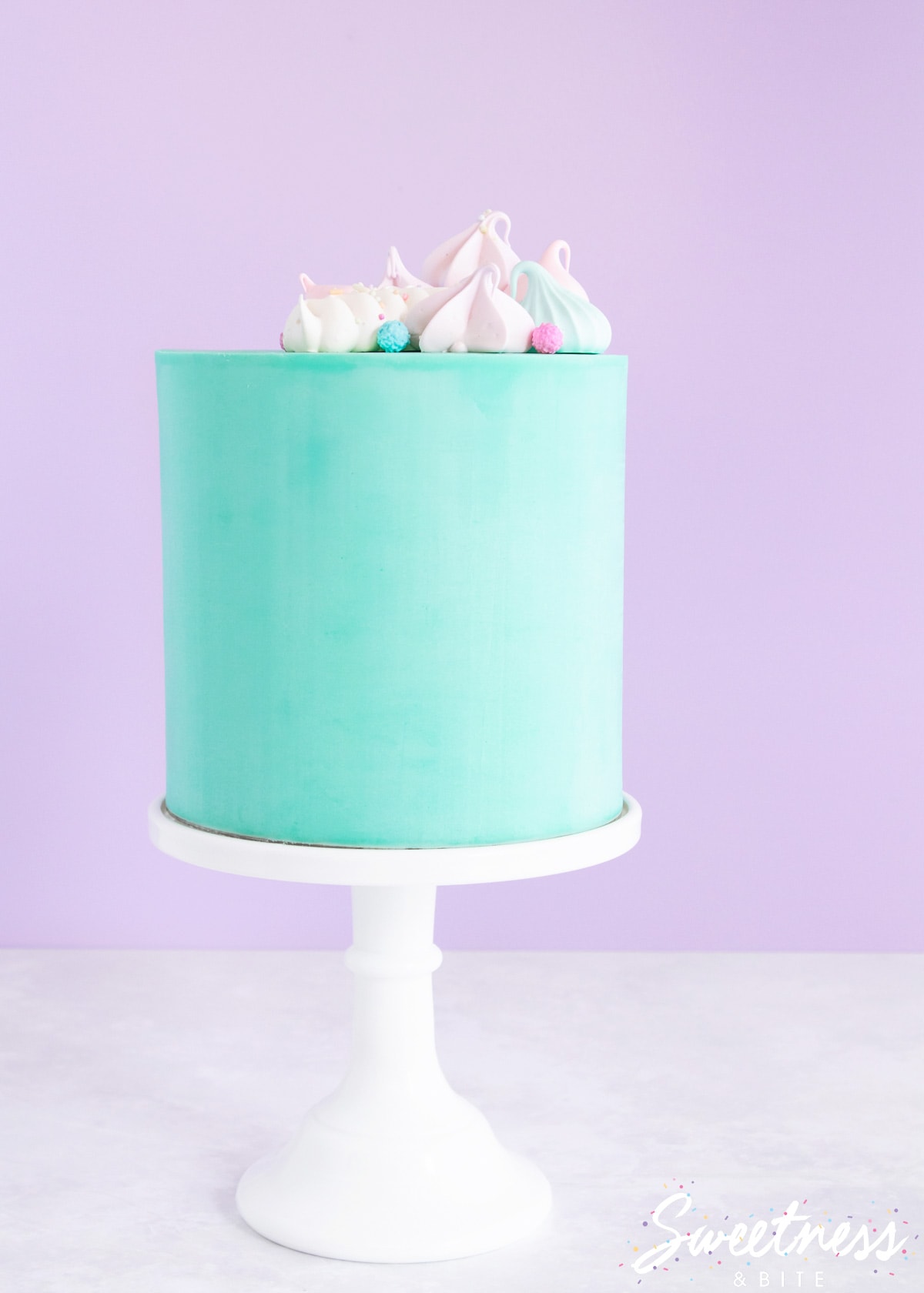 A cake covered in teal ganache on a white cake stand, topped with pastel coloured meringues.
