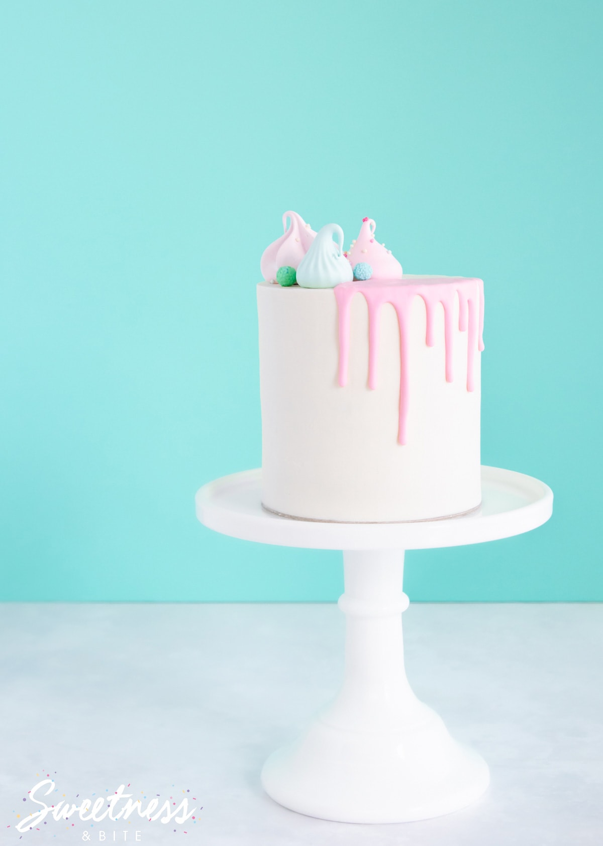 A cake covered in white chocolate ganache with a pink coloured ganache drip, on a white cake stand.