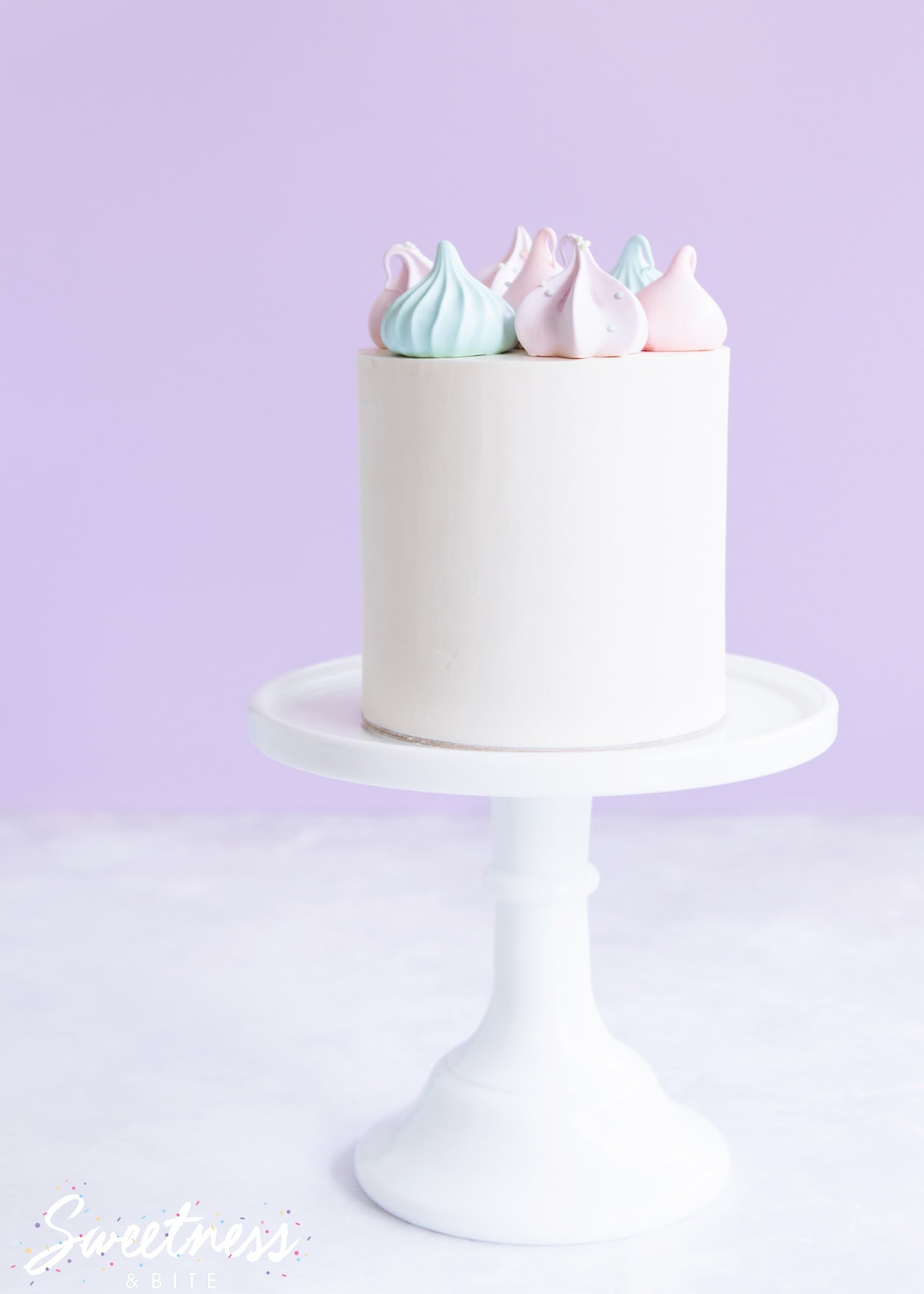 A cake covered in whitened white chocolate ganache on a white cake stand decorated with pastel meringues.