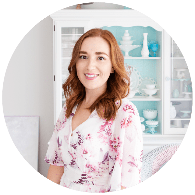 A photo of Natalie, a rather pale redhead in a floral jumpsuit, standing in front of a white-painted hutch dresser displaying cake stands.