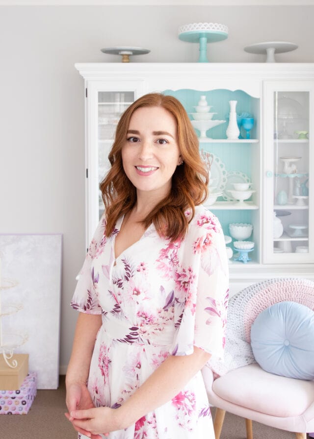 A photo of Natalie, a rather pale redhead in a floral jumpsuit, standing in front of a white-painted hutch dresser displaying cake stands.