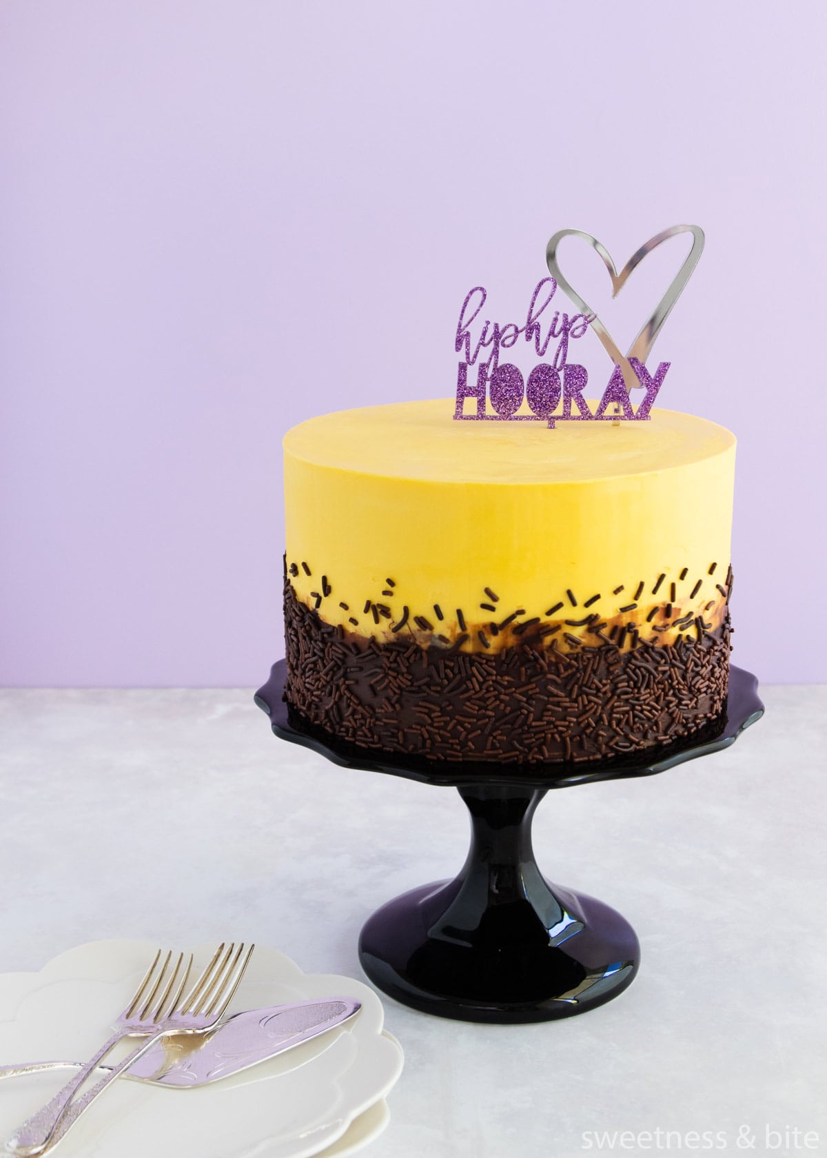 A cake with yellow and dark chocolate ganache with chocolate sprinkles and a purple 