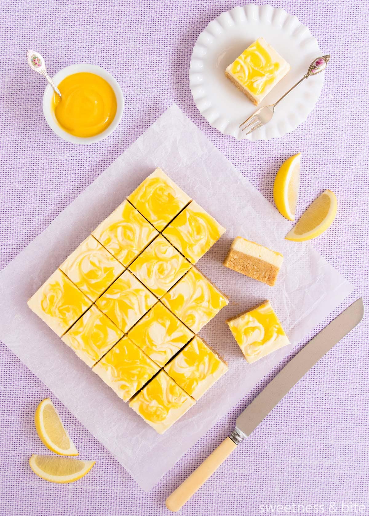 The slice, cut into squares on a pale purple background, with a knife and a bowl of lemon curd.