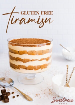 Gluten free tiramisu in a footed glass trifle bowl, on a grey/blue background, with small white bowls and a serving spoon.
