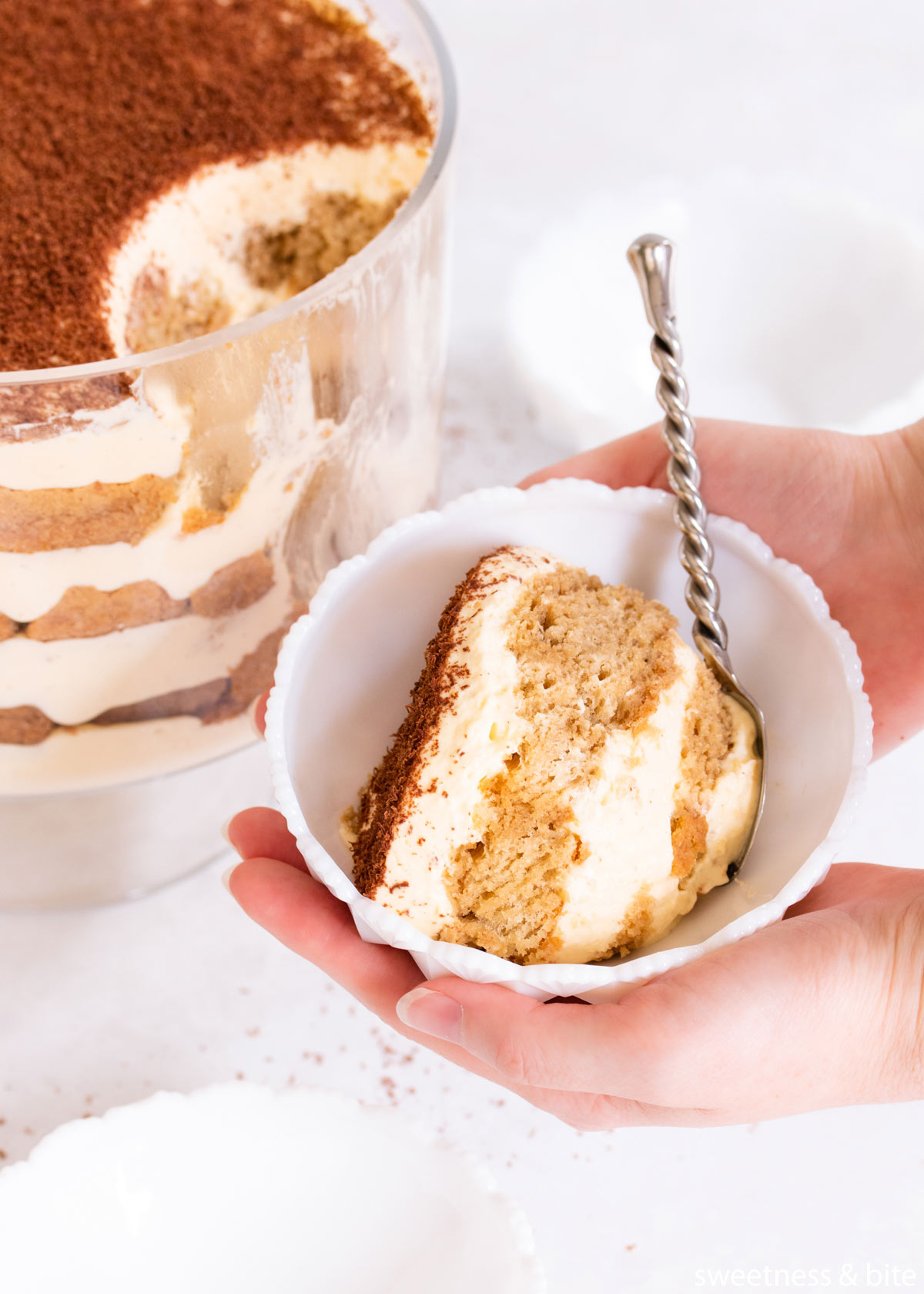 Close-up of two hands holding a bowl of tiramisu in a white milk glass bowl.