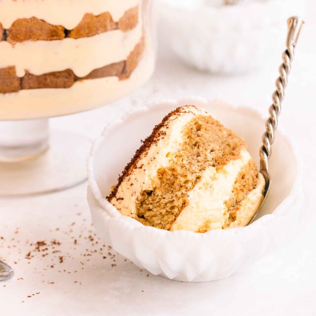 Gluten free tiramisu in a footed glass trifle bowl, on a grey/blue background, with small white bowls and a serving spoon.