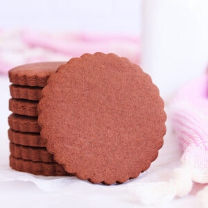 A stack of scalloped-edge round gluten free chocolate cookies, with one cookie resting on its side against the stack.