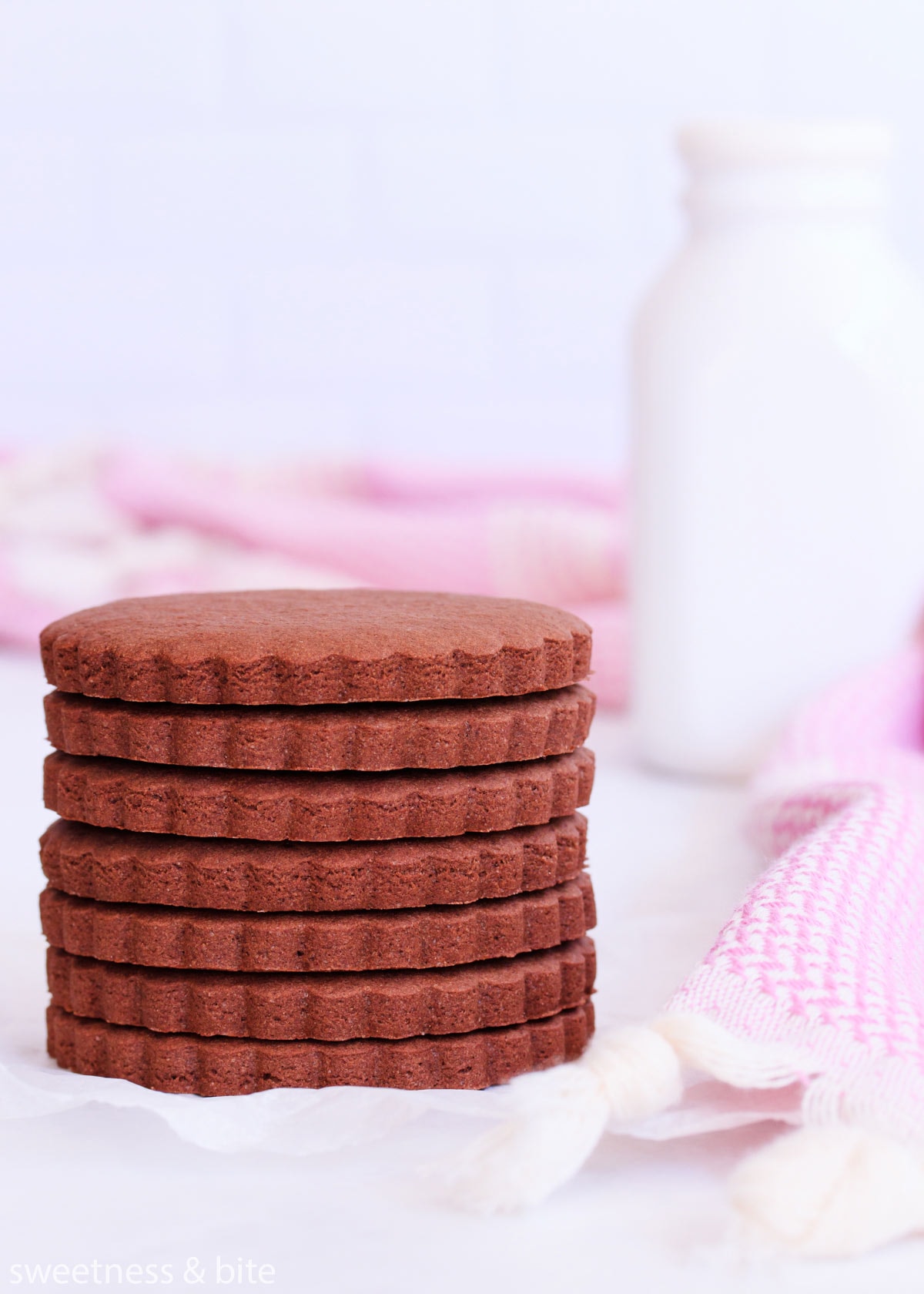 A stack of scalloped-edge round gluten free chocolate cookies, with a purple cloth and a white milk bottle.