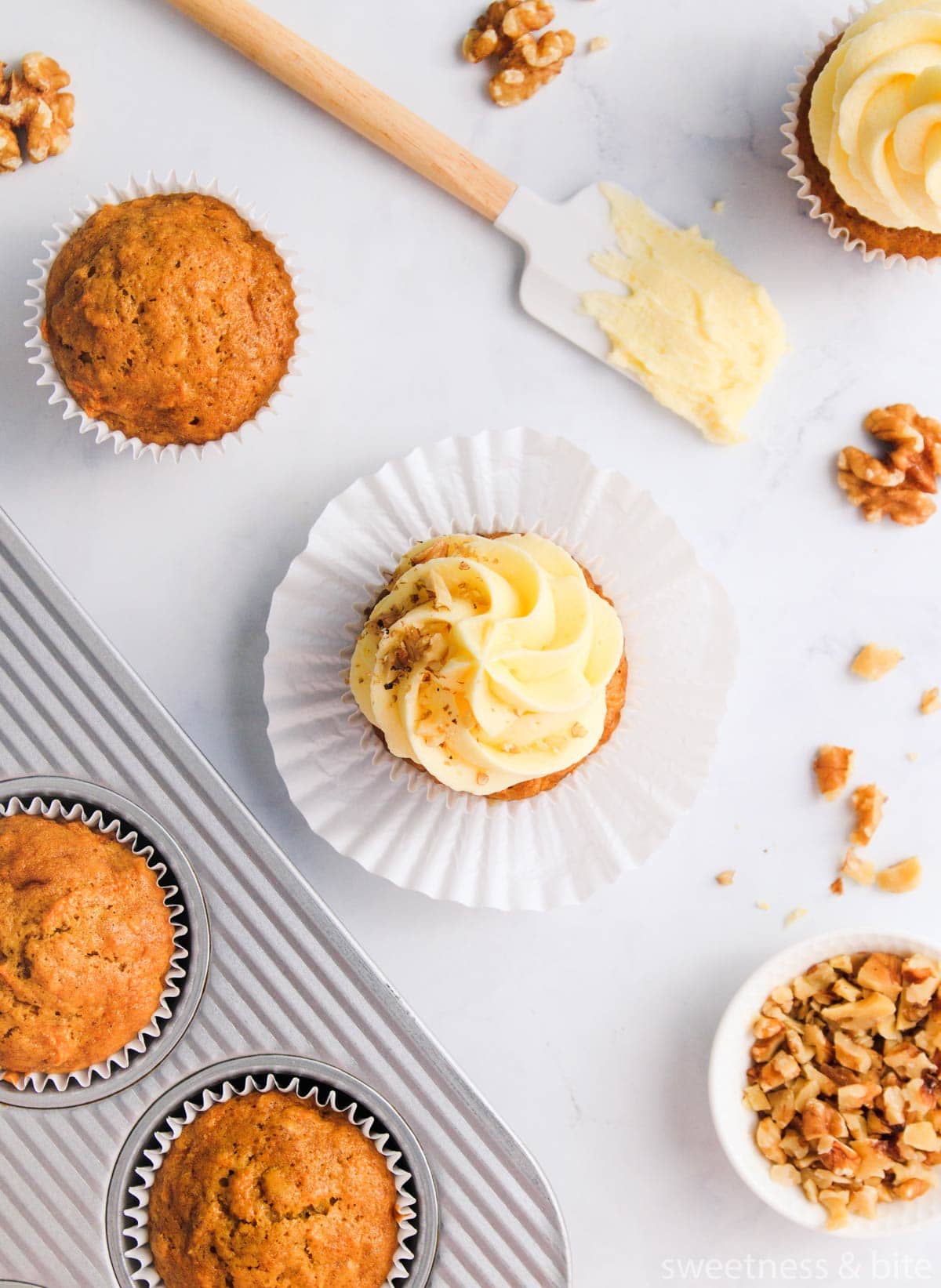 A flat-lay showing several carrot cupcakes, some iced and some uniced, with scattered walnuts and a spatula spread with cream cheese icing, on a grey marble background.
