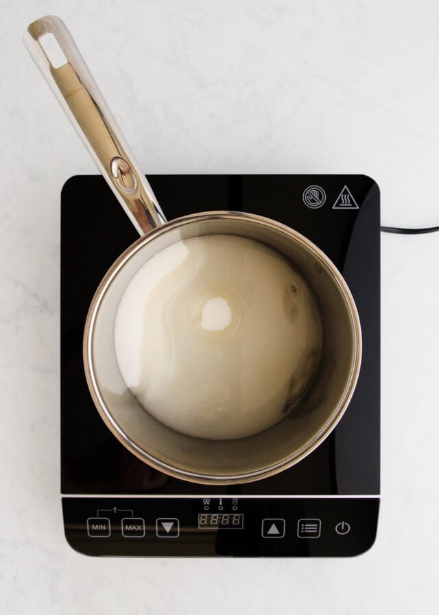 The sugar dissolving in a stainless steel saucepan on a small black induction cooker.
