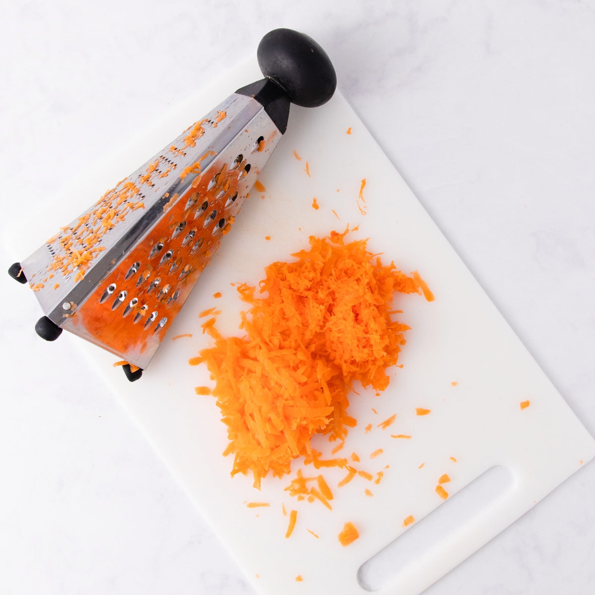 Shredded carrots on a white chopping board, next to a pyramid box grater.