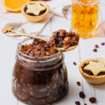A large glass jar of fruit mince, on a white wooden background, with fruit mince pies and a glass jug of brandy in the background.