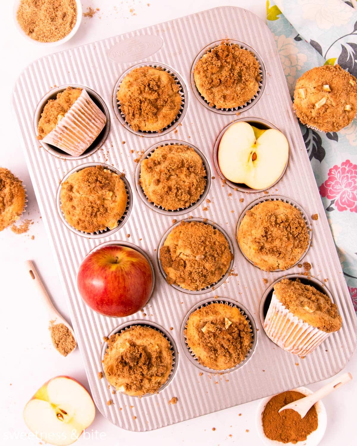 Gluten free apple muffins in a metal muffin pan, with a red apple and small dish of cinnamon streusel.