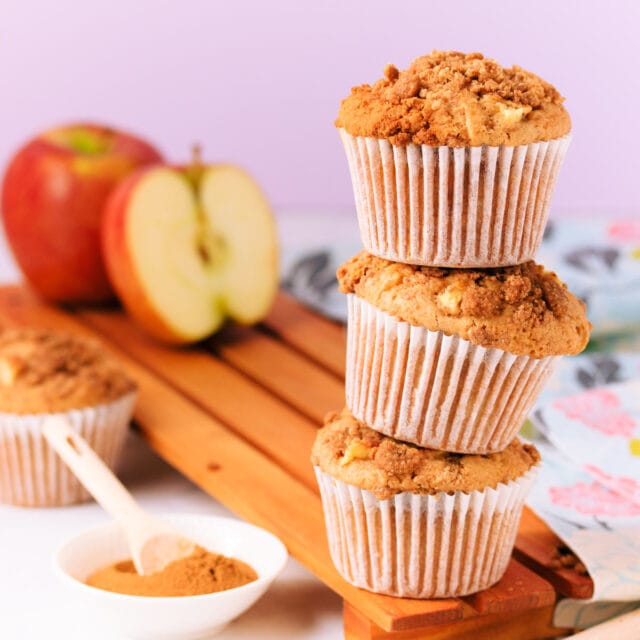 Three gluten free apple muffins stacked on top of each other, on a wooden board, with an apple and small dish of cinnamon in the background.