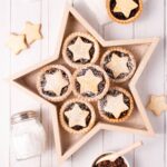 Gluten free mince pies served in a star-shaped wooden box, on a white wooden background, with a small bowl of fruit mince and shaker of icing sugar.