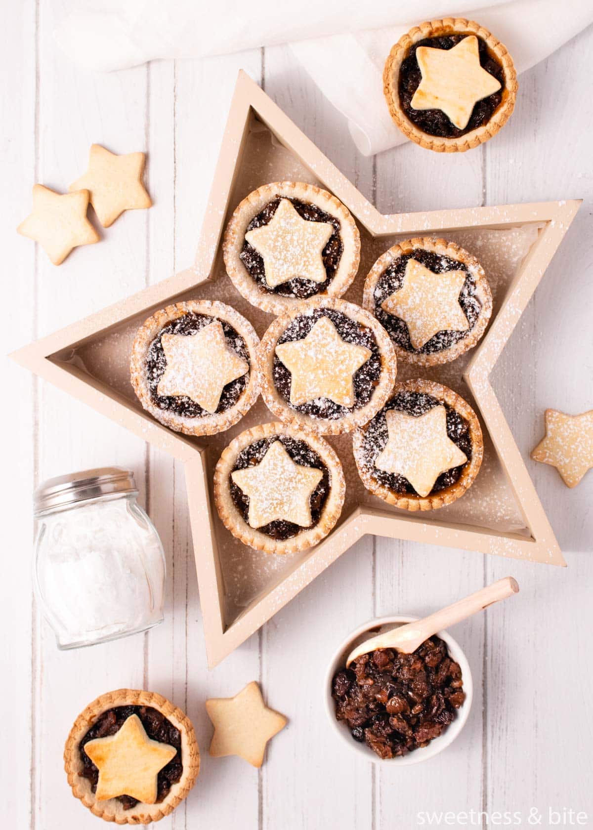 Gluten free mince pies served in a star-shaped wooden box, on a white wooden background, with a small bowl of fruit mince and shaker of icing sugar.