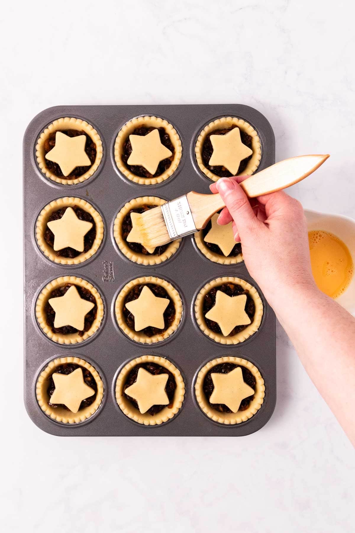 A hand holding a pastry brush, brushing an egg wash on the tops of the mince pies.