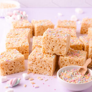 A stack of gluten free rice krispie treats on a pale purple background with a small bowl of pastel sprinkles.