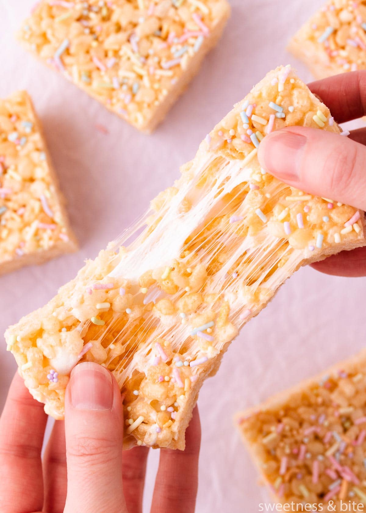 A gluten free rice krispie treat being pulled apart by two hands, showing the marshmallow stretching as it is pulled.