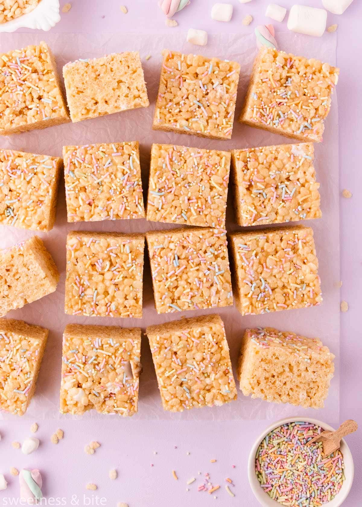 Squares of rice krispie treat arranged haphazardly on a sheet of crinkled parchment on a lilac background, with mini marshmallows, rice krispies and sprinkles scattered around. 