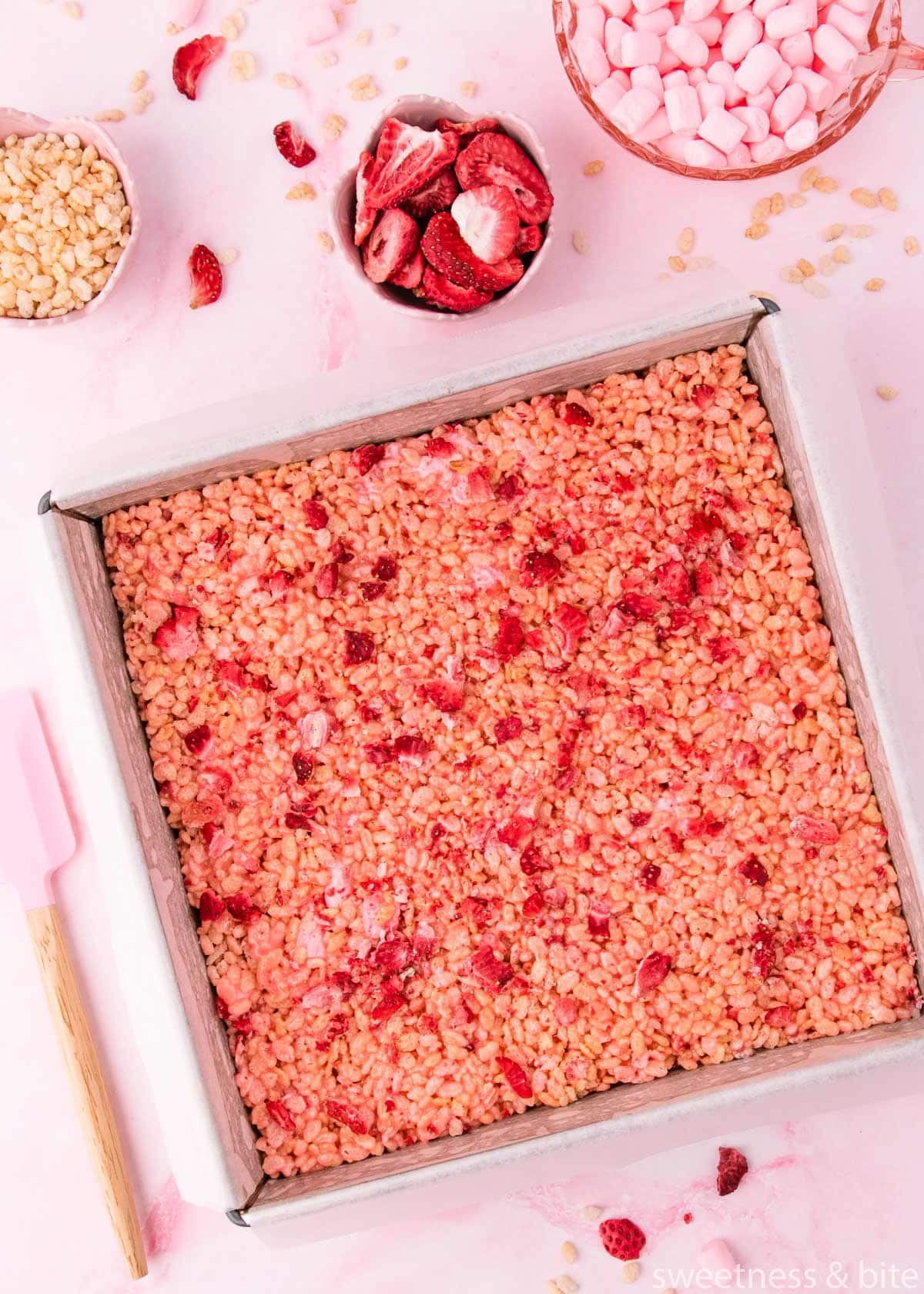 A square pan of strawberry rice krispie treats on a pink marble background with a pink spatula and scattered freeze-dried strawberries.